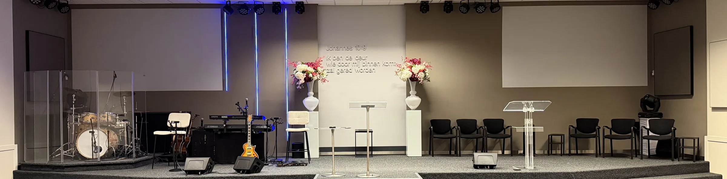 Het podium in de kerkzaal van De Deur Amersfoort, met muziekinstrumenten, bloemen en op de wand de tekst uit Johannes 10:9: Ik ben de deur, wie door Mij binnenkomt zal gered worden