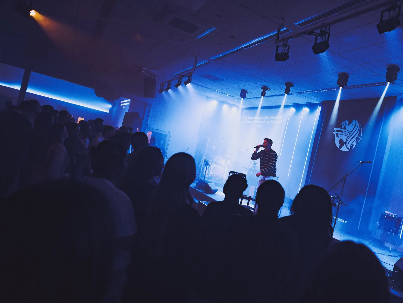 Een volle zaal tijdens een dienst bij De Deur, met mensen die meezingen voor het podium